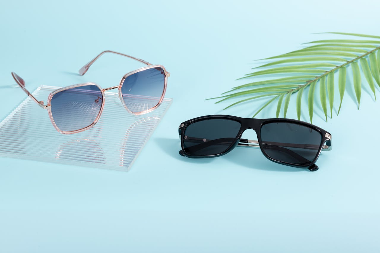 Services-02 Two pairs of trendy sunglasses on a pastel surface with palm leaf decor.